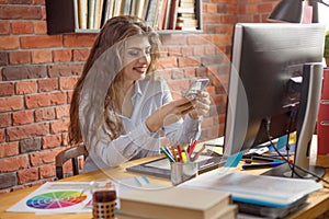 Young female with long hair working at home or in a loft style office. She chatting via smartphone and smiling. Designer, editor,