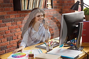 Young female with long hair working at home or in a loft style office. She speaking via smartphone and smiling. Designer, editor,