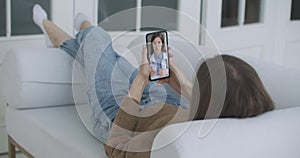 Young female lying on the couch at Home Using Smartphone to Talk to Her Doctor via Video Conference Medical App. Woman