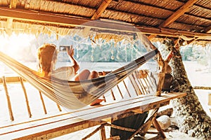 Young female lying in hammock and taking a landscape photo using modern smartphone on Samui island in Thailand. Careless