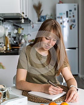 Young female making notes in notebook sitting in kitchen. Planning buying. Making list of products