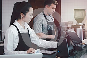 Young female and male  barista wearing apron standing behind coffee shop cashier counter, waitress and waiter staff working at