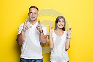 Young female and male closing thier eyes crossing fingers with hope while anticipating important news standing against yellow