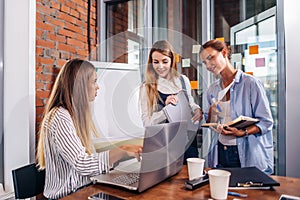 Young female manager sitting at desk pointing at laptop explaining giving tasks to her employees standing writing the