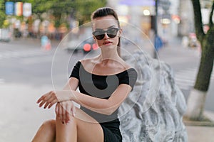 Young female model posing in the city with attitude, in a black dress with round sunglasses, over blurred background.