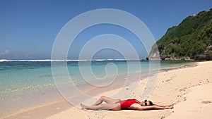 Young female model in red swimsuit lying near water on wild tropic sand beach and drinking coconut juice, Bali