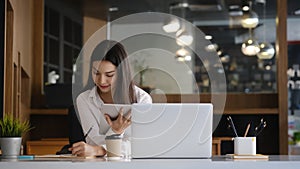 Young female office worker holding digital table and writing information on paper at office.