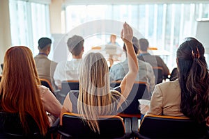 A young female participant is raising a hand to ask a question during a business lecture in the conference room. Business, people