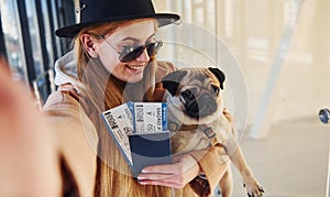Young female passenger in warm clothes holding tickets and cute dog in hands in airport hall