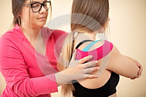 Young female patient wearing kinesio tape on her shoulder exercising with a professional physical therapist. Kinesiology, physio.