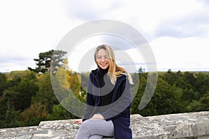 Young female person sitting on concrete railing with tree background.