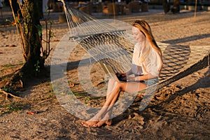 Young female person using laptop and sitting on sand in wicker hammock.