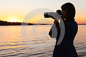 Young female photographer taking photo of riverside sunset with professional camera