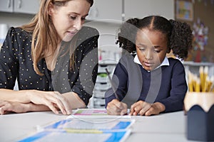 Young female primary school teacher working one on one with a schoolgirl watching her write at a table in a classroom, close up