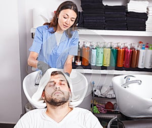 Young female professional washing cheerful male's hair in salon