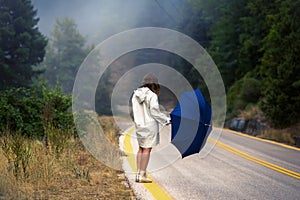 Young female in a raincoat and umbrella on the road in the fog. Travel of women in the raincoat hitchhiking in the rain