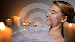 Young female relaxing in warm bath with foam bubbles and candles, evening