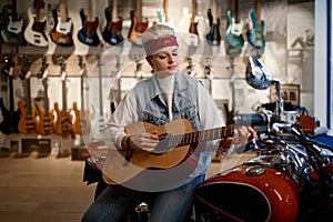 Young female rocker playing guitar at music shop sitting on motorcycle