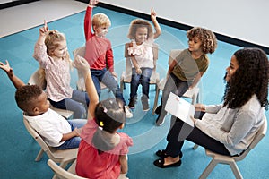 Young female school teacher reading a book to infant school children, sitting on chairs in a circle in the classroom raising hands