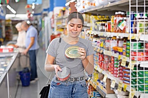 Young female shopper choosing canned food in supermarket