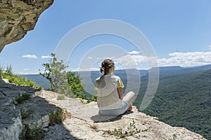 Young female sit on cliff on peak in high mountains looking over green mountains with feeling of relexation.