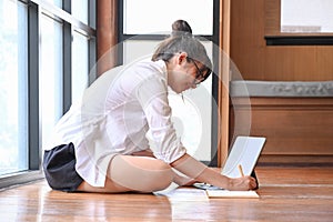 Young female sitting on floor near window using computer tablet and making note on notebook.