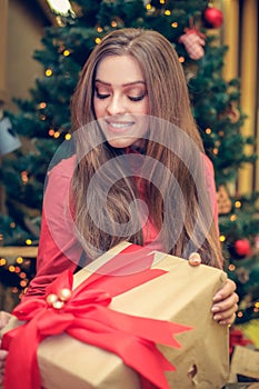 Young female is sitting in front of the christmas tree and have gift box with red ribbon in her hands