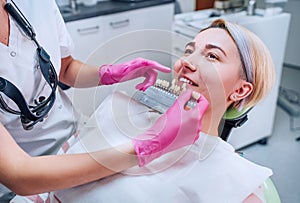 Young female sitting in stomatology clinic chair and smiling showing her teeth. Dentist doctor choosing tooth colors with shade photo