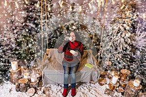 Young female sitting on a swing with a blanket under the flashlights and drinkig a cup of coffee in a snow-covered park