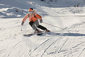 A young female skier in an orange jacket turns carving arches on the slope. A beautiful sunny day in the mountains, on the ski