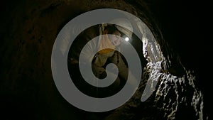 Young female speleologist climbing into the dark narrow cave explores the cave with flashlight