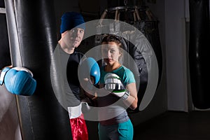 Young female spoty boxer in boxing gloves standing with her trainer and boxing punching bag