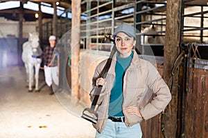 Young female stable worker carrying pitchfork to stack hay for horses
