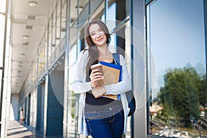 Young female IT student, with books and backpack. Staying outside before lesson and drinking coffee