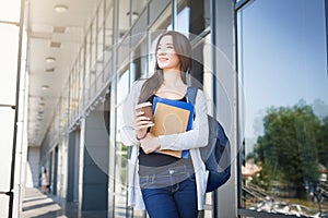 Young female IT student, with books and backpack. Staying outside before lesson and drinking coffee