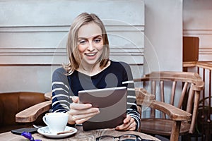 Young female student is chatting on digital tablet with friend while sitting in cafe, surprised attractive woman using