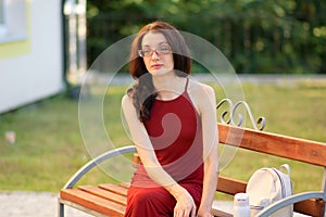 Young Female Student in Eyesglasses is Sitting on the Bench During Sunny Day in the Summer.