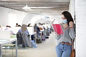 Young female student with face mask reading a book inside a classroom. Presential classes at the university