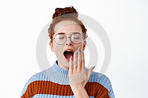 Young female student with ginger hair combed in hairbun, wearing glasses, yawning with opened mouth, covering it with