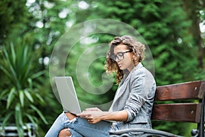 Young female student sitting on a bench in park, she is working on laptop