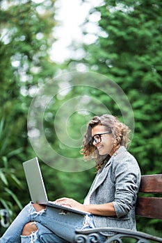 Young female student sitting on a bench in park, she is working on laptop