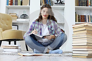 Young female student studying at home, sitting on floor against cozy domestic interior, surrounded with pile of books