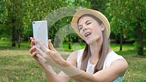 Young female student in white shirt sitting on grass on college campus doing selfie on mobile phone