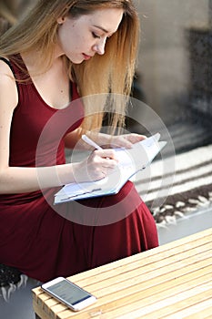 Young female student writing down in notebook at university, smartphone on table.