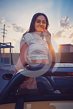 Young female in sunglasses and white t-shirt is eating pizza while posing standing in yellow car cabriolet. Fast food