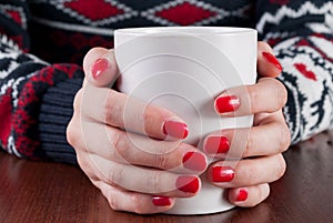 Young girl in sweater holding white coffee cup in hand with red nails on wooden desk