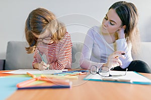 Young female teacher giving private lesson to child, little girl sitting at her desk writing in notebook