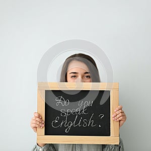 Young female teacher holding chalkboard with words DO YOU SPEAK ENGLISH? on light background. Space for text