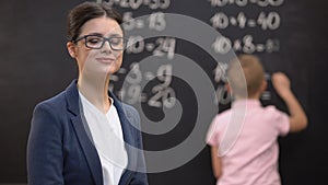 Young female teacher looking to camera, schoolboy solving math exercises, job