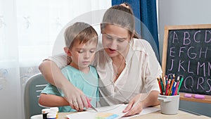 Young female teacher showing her little student how to use scissors. Boy cutting paper with scissors while doing